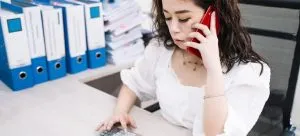 A woman in an office using a calculator while talking on a phone.